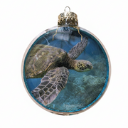 A round clear glass Christmas ornament featuring a printed sea turtle swimming underwater scene, with a glittery blue rim and a gold hanging cap on a white background.