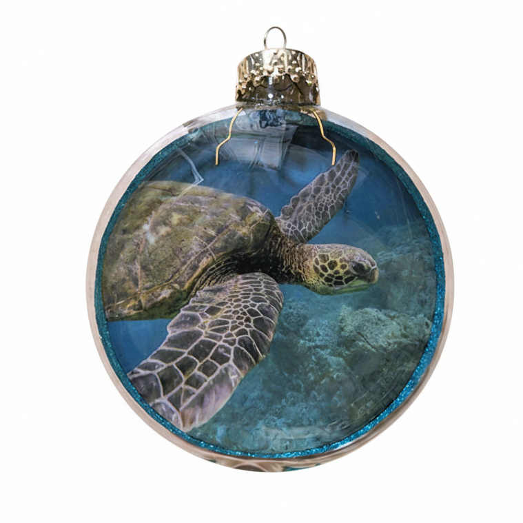 A round clear glass Christmas ornament featuring a printed sea turtle swimming underwater scene, with a glittery blue rim and a gold hanging cap on a white background.