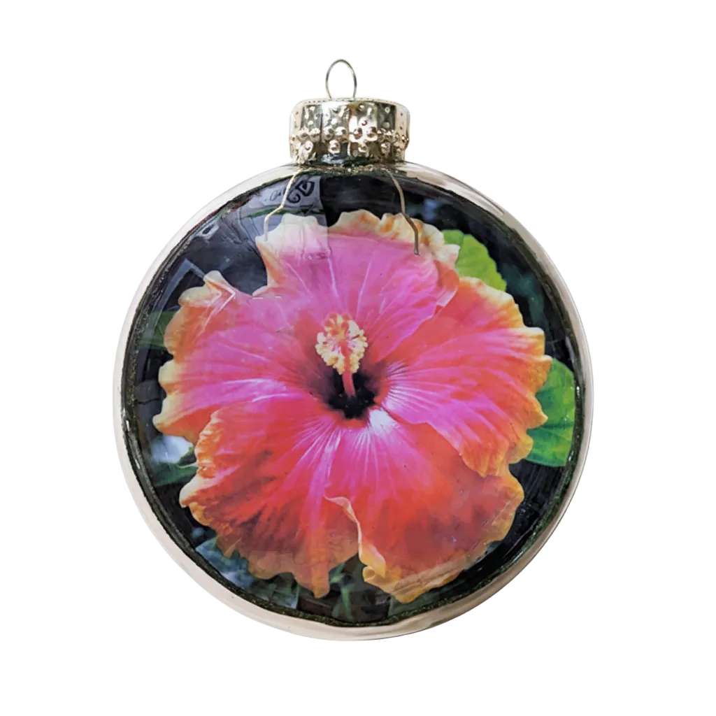A round glass Christmas ornament featuring a vibrant pink hibiscus flower with orange-yellow edges and green leaves, complete with a silver glittery cap and hanging loop, set against a plain white background.