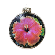 A round glass Christmas ornament featuring a vibrant pink hibiscus flower with orange-yellow edges and green leaves, complete with a silver glittery cap and hanging loop, set against a plain white background.