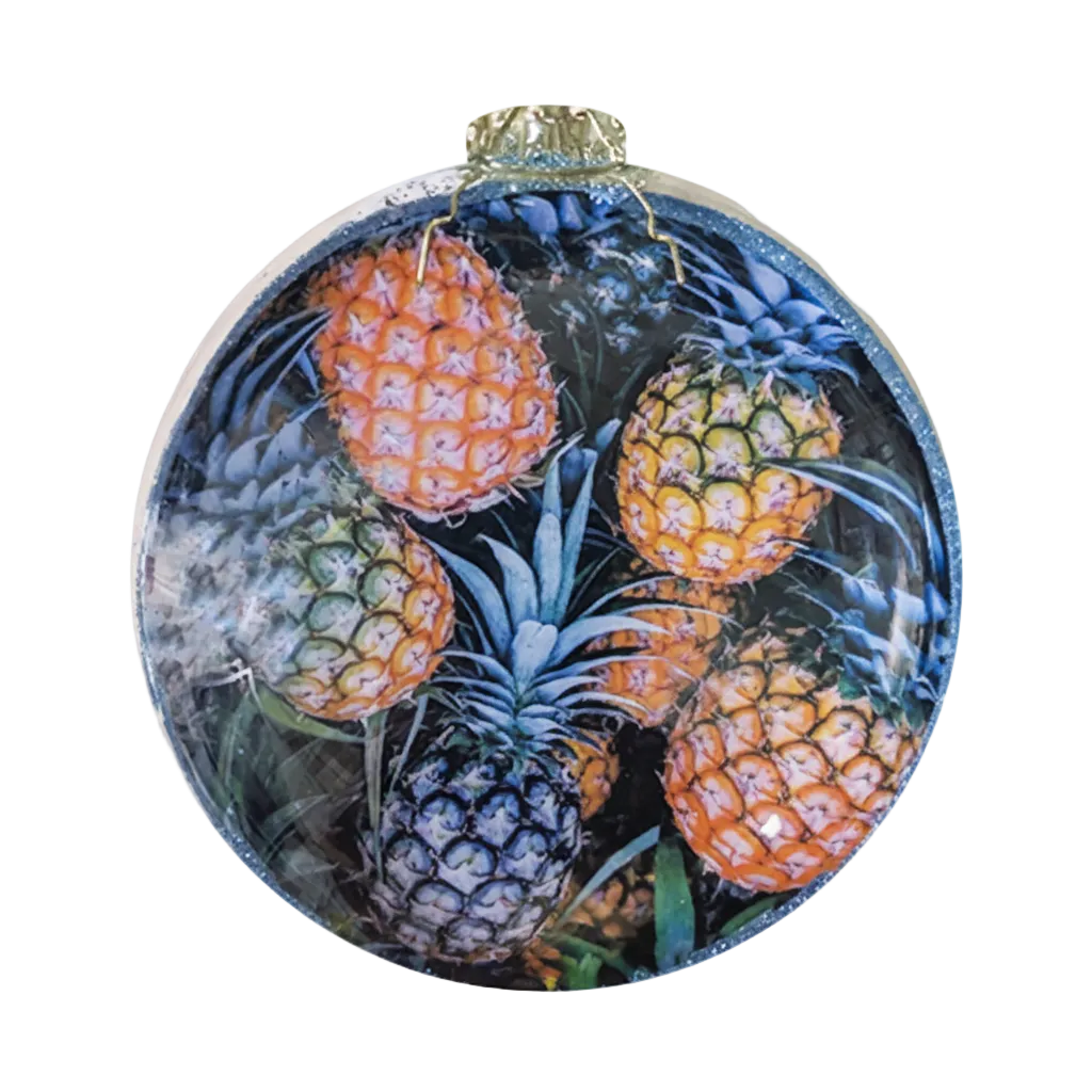 A round glass Christmas ornament featuring a colorful print of pineapples on a dark background, complete with a gold hanging loop at the top.