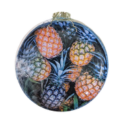 A round glass Christmas ornament featuring a colorful print of pineapples on a dark background, complete with a gold hanging loop at the top.