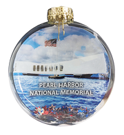 A round clear glass ornament featuring a printed image of the Pearl Harbor National Memorial, with blue sky and water, a white ship, an American flag, and a gold hanging cap at the top.