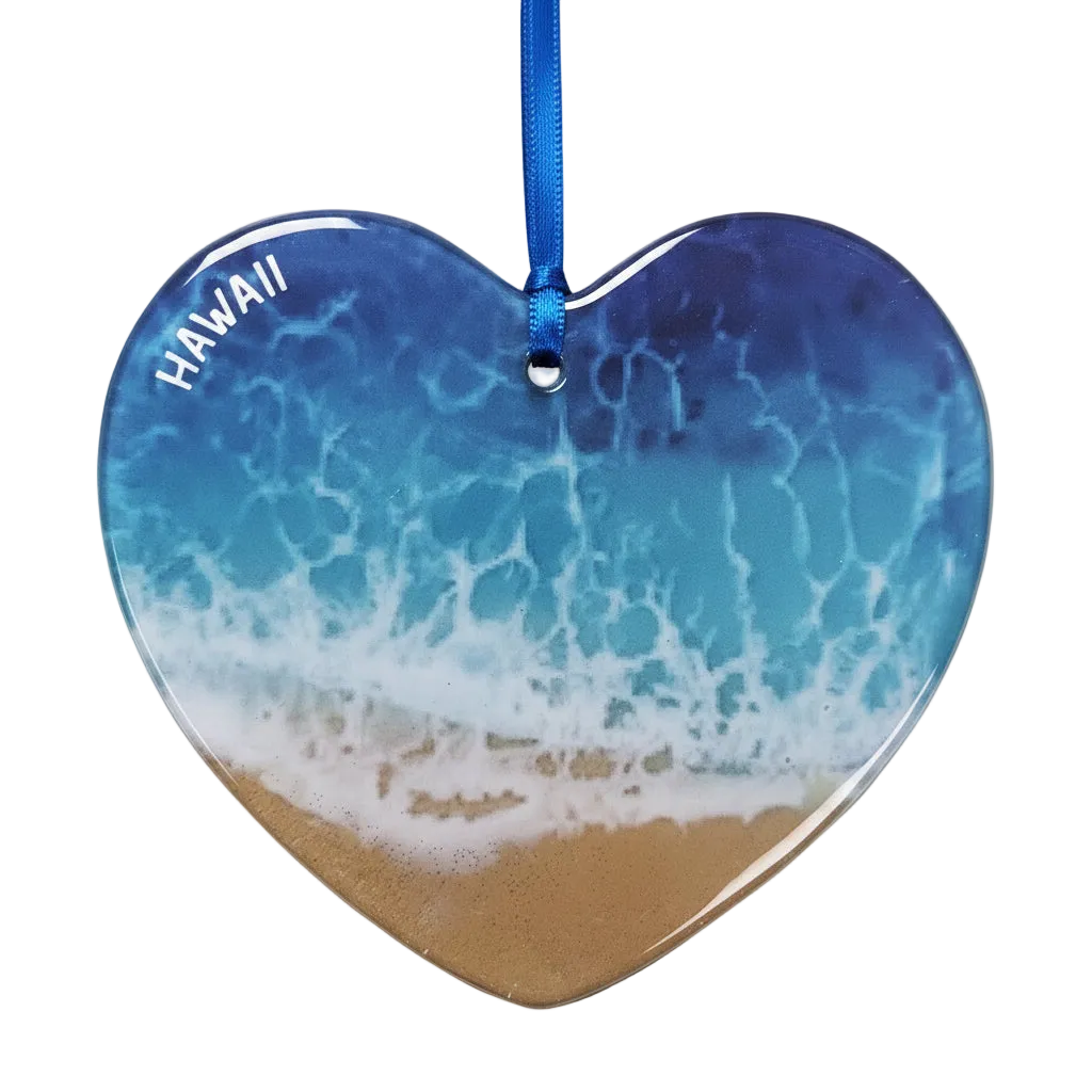 Heart-shaped Hawaii beach ornament featuring a blue and white ocean wave pattern with tan sand at the bottom and the word 'HAWAII' printed in white along the upper left edge, hanging from a blue ribbon on a white background.