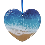 Heart-shaped Hawaii beach ornament featuring a blue and white ocean wave pattern with tan sand at the bottom and the word 'HAWAII' printed in white along the upper left edge, hanging from a blue ribbon on a white background.