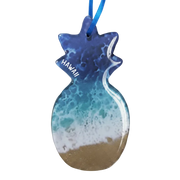 A pineapple-shaped resin ornament featuring an ocean wave design with blue and teal water, white foam, and a sandy beige bottom. The word 'HAWAII' is printed in white on the side, and a blue ribbon is attached at the top, shown against a plain white background.
