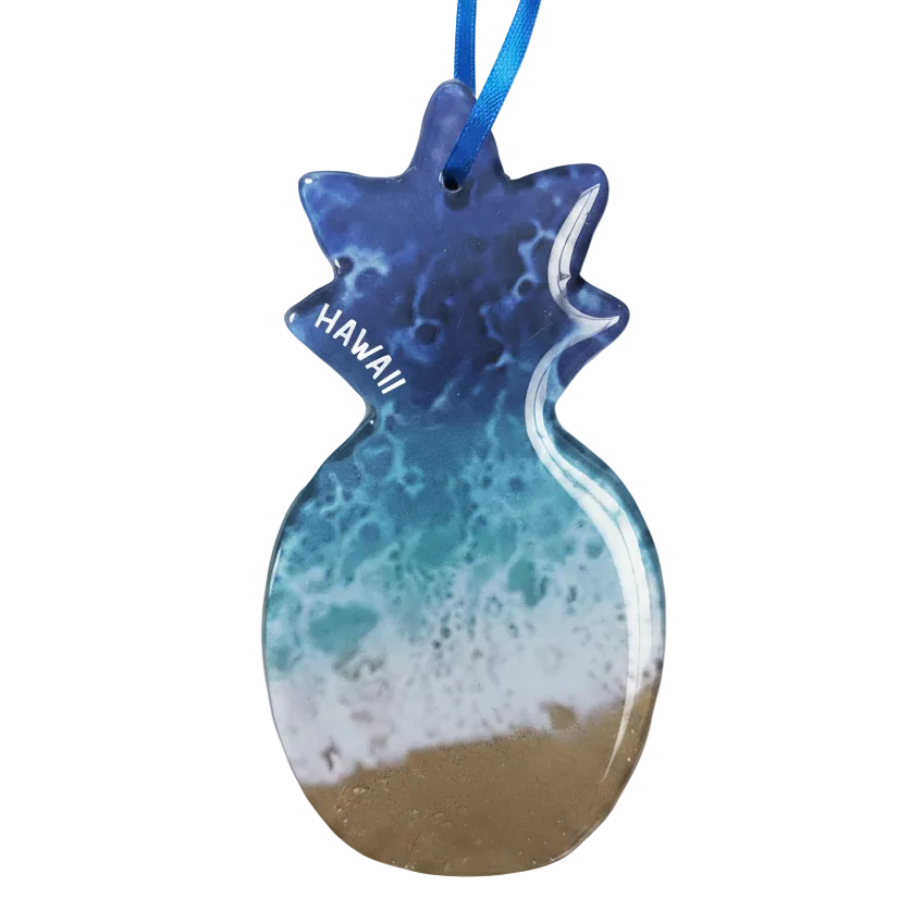 A pineapple-shaped resin ornament featuring an ocean wave design with blue and teal water, white foam, and a sandy beige bottom. The word 'HAWAII' is printed in white on the side, and a blue ribbon is attached at the top, shown against a plain white background.
