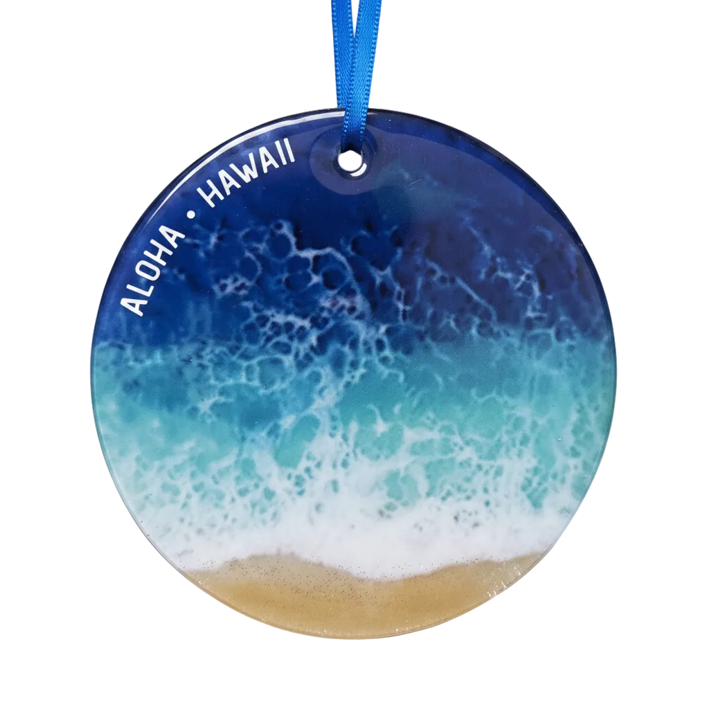 Round glass ornament featuring an ocean wave pattern with white foam and a sandy beach at the bottom, with 'ALOHA • HAWAII' printed in white along the left side. The ornament hangs from a blue ribbon on a white background.