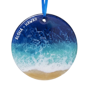 Round glass ornament featuring an ocean wave pattern with white foam and a sandy beach at the bottom, with 'ALOHA • HAWAII' printed in white along the left side. The ornament hangs from a blue ribbon on a white background.