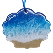 A scalloped shell-shaped ornament featuring a blue and white ocean wave pattern with white 'HAWAII' text and a glittery gold sand base, hanging from a blue ribbon on a white background.