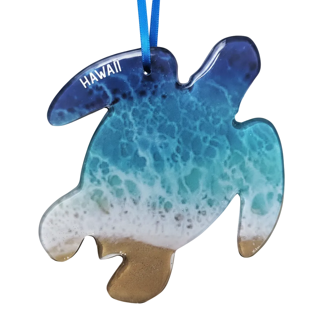 A turtle-shaped Hawaiian Christmas ornament with a blue and turquoise ocean wave pattern, white foam, and a sandy beige bottom, hanging from a blue ribbon. The word 'HAWAII' is printed in white on the left side.