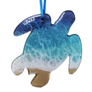 A turtle-shaped Hawaiian Christmas ornament with a blue and turquoise ocean wave pattern, white foam, and a sandy beige bottom, hanging from a blue ribbon. The word 'HAWAII' is printed in white on the left side.